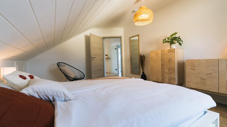 Ein gemütliches Schlafzimmer mit einem großen Bett und einer modernen Einrichtung. Die Wände sind hell, und ein Holzregal sowie Pflanzen sorgen für eine angenehme Atmosphäre.