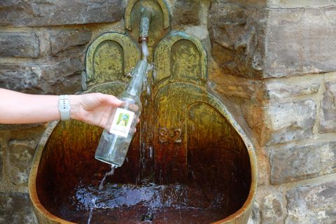 Eine Hand hält eine Glasflasche unter einem alten Wasserbrunnen. Wasser fließt aus dem Brunnen in die Flasche.