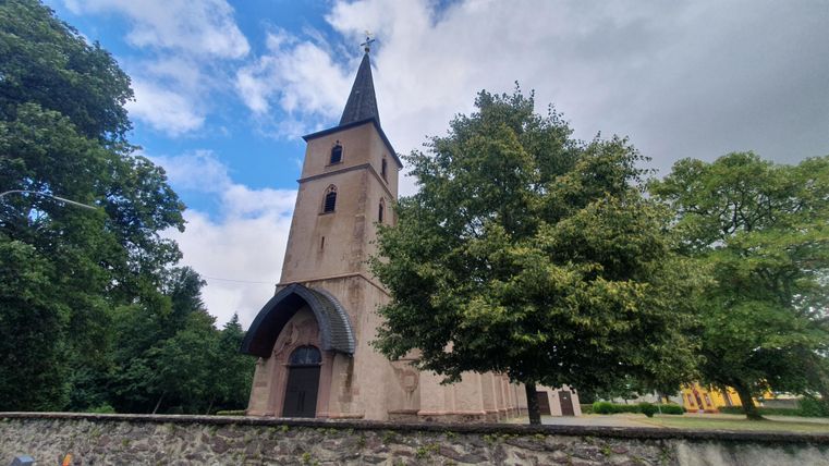 Eine alte Kirche mit einem hohen Turm und klassischen architektonischen Details. Umgeben von Bäumen und einem klaren Himmel.