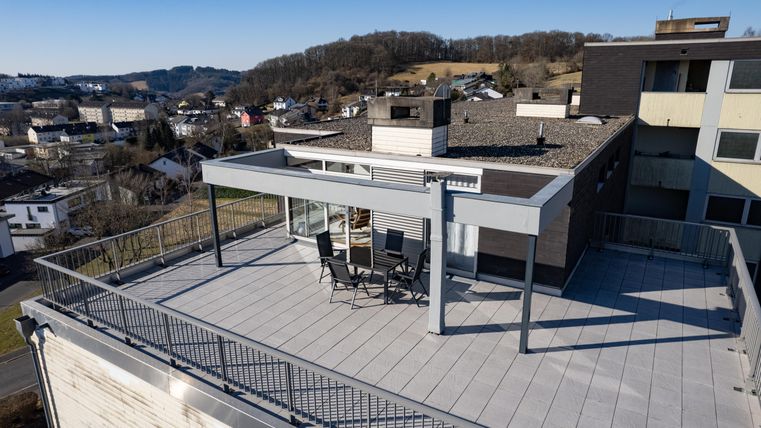Eine moderne Dachterrasse mit Ausblick auf eine Stadtlandschaft. Auf der Terrasse stehen Stühle und ein Tisch unter einem Überdach.