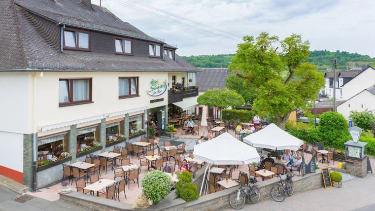 A cozy café with a terrace and many outdoor tables. In the background, trees and a peaceful environment can be seen.
