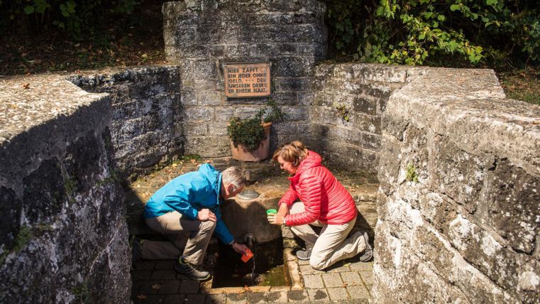 Zwei Personen beugen sich über eine Wasserquelle, die von steinernen Wänden umgeben ist. Im Hintergrund ist ein Schild mit Informationen über die Quelle zu sehen.