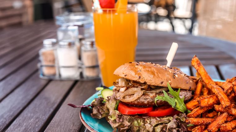 Ein saftiger Burger mit Salat, Tomate und Zwiebeln, serviert mit süßen Kartoffelpommes. Dazu ein erfrischendes Getränk in einem Glas.