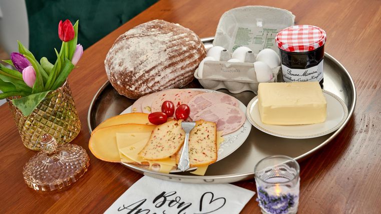 Ein Frühstücksgericht auf einem Tablett mit Brot, Käse, Schinken, Tomaten und Eier. Dazu sind eine Blume und eine Kerze auf dem Tisch platziert.