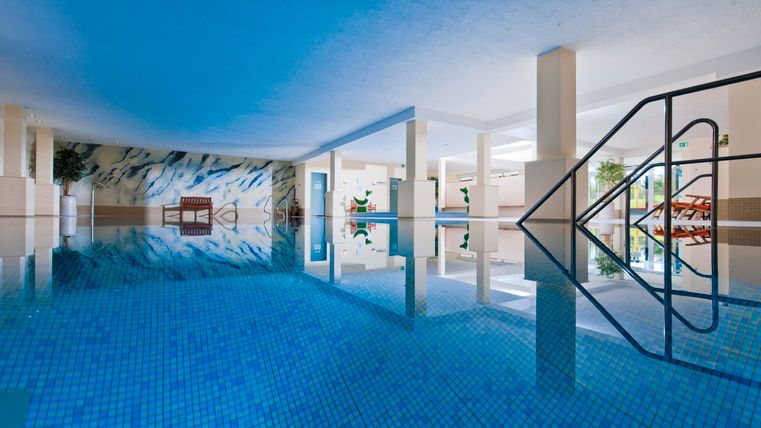 Ein modernes Hallenbad mit blauem Wasser und einer hellen, einladenden Atmosphäre. Umgeben von Pflanzen und mit einem Blick auf bequeme Liegen.