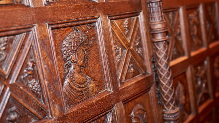 Eine kunstvoll geschnitzte Holzwand mit detaillierten Reliefs von Figuren. Die Schnitzereien zeigen historische oder mythologische Motive.