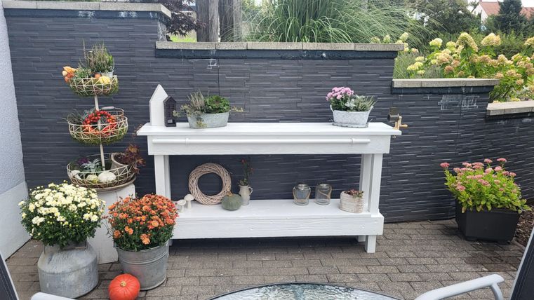 Ein schöner Gartentisch mit mehreren Pflanzenutteln und Töpfen. Umgeben von bunten Blumen und einer modernen Mauer.