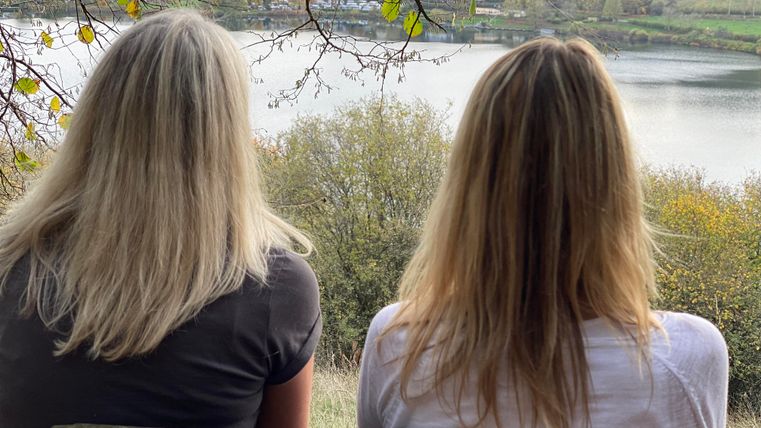 Zwei Frauen sitzen auf einer Bank und blicken auf einen ruhigen See. Im Hintergrund sind Bäume und Hügel zu sehen.