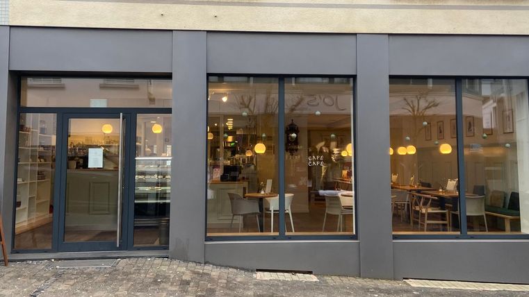 Ein modernes Café mit großen Fenstern und einem einladenden Innenraum. Draußen steht ein Schild, das Gäste anlockt.