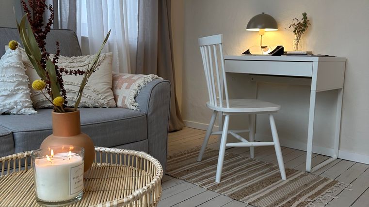 Ein gemütliches Wohnzimmer mit einem grauen Sofa, einem weißen Schreibtisch und einem Stuhl. Ein Tisch mit einer Kerze und einer Pflanze ergänzt die entspannte Atmosphäre.