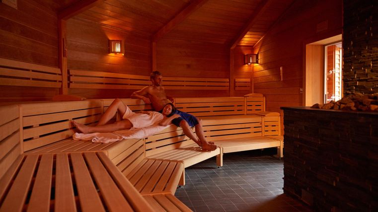Ein gemütlicher Sauna-Raum mit Holzregalen und sanfter Beleuchtung. Zwei Menschen entspannen sich auf den Bänken.