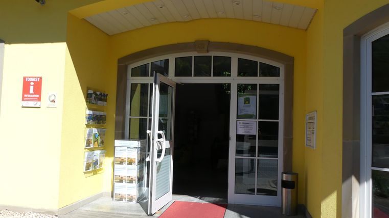 Ein Eingang zu einem Gebäude mit gelber Fassade und einer offenen Glastür. Roter Teppich und Informationsmaterial stehen bereit.