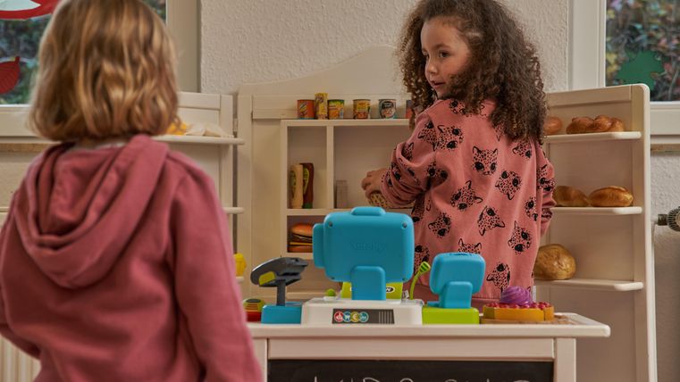 Ein kleiner Spielbereich mit zwei Kindern, die an einem Spielwarenstand spielen. Das Kind rechts steht hinter dem Stand und sortiert Lebensmittel, während das andere Kind zuschaut.