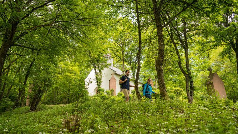 Two hikers walk through a forest with a small white chapel in the background.