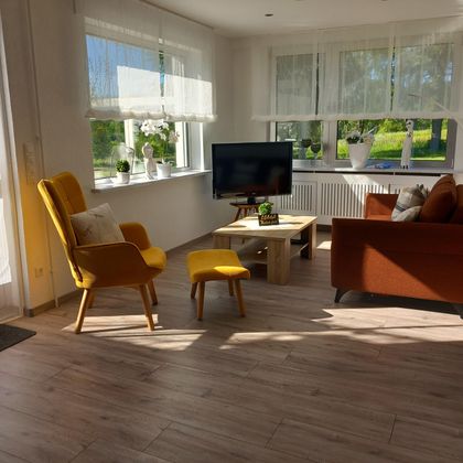 A bright living space with large windows and plenty of natural light. There is a television, a brown sofa, and a yellow armchair.