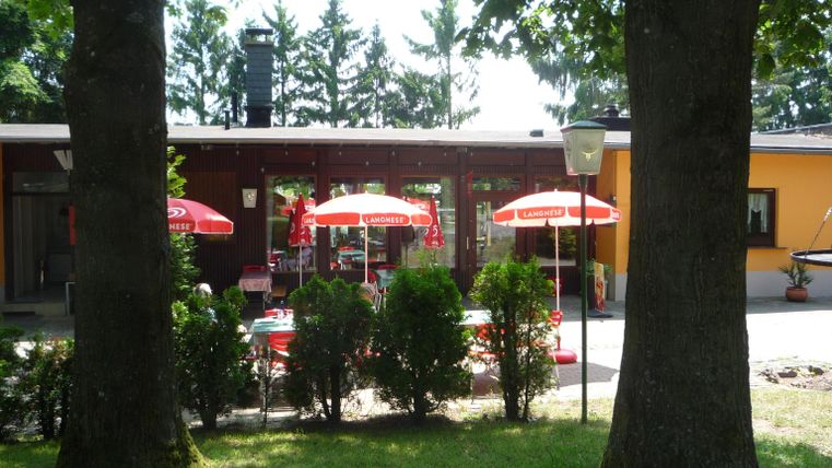 Ein gemütliches Café mit roten Sonnenschirmen im Freien. Umgeben von Bäumen und einem grünen Rasen.