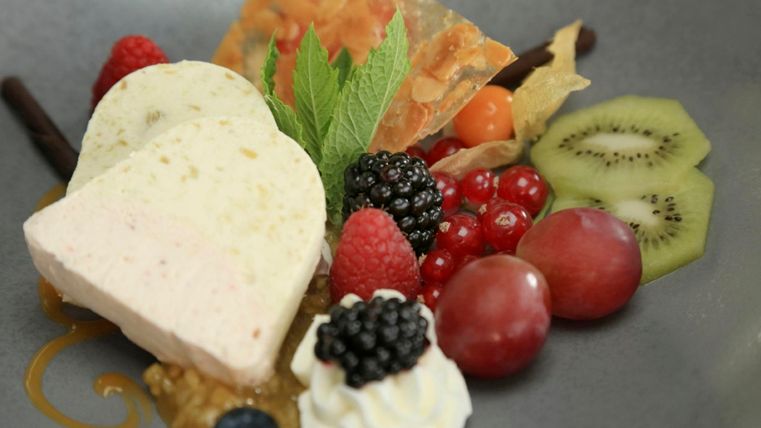 Ein kunstvoll angerichtetes Dessert mit einer Herzform aus Eis, frischen Beeren und fruchtigen Garnituren. Grünes Obst und eine dekorative Süßspeise ergänzen das Gericht.