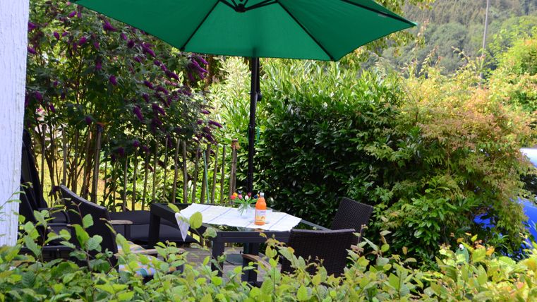 Ein gemütlicher Sitzbereich im Garten mit einem grünen Sonnenschirm. Der Tisch ist für eine kleine Erfrischung gedeckt, umgeben von üppigem Grün.