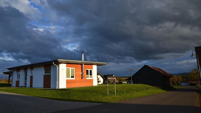 Ein modernes Haus mit roter Fassade steht in einer ländlichen Umgebung. Dunkle Wolken ziehen am Himmel vorbei.