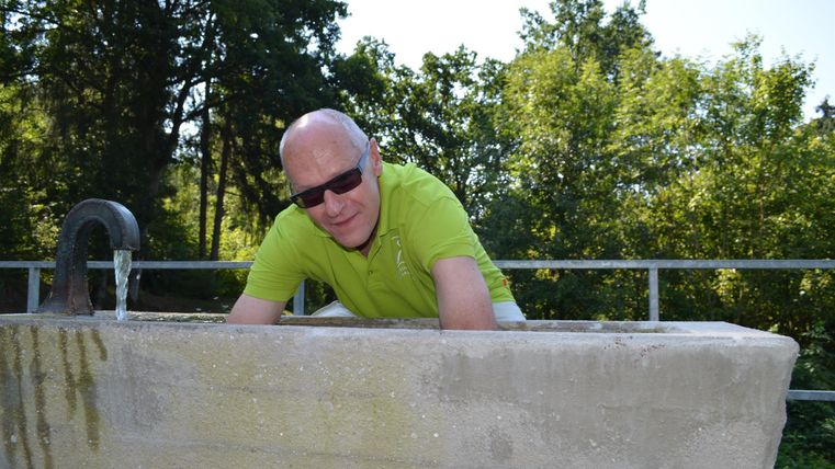Ein Mann in einem hellgrünen Shirt beugt sich über eine Betonkante. Im Hintergrund sind grüne Bäume und ein blauer Himmel zu sehen.