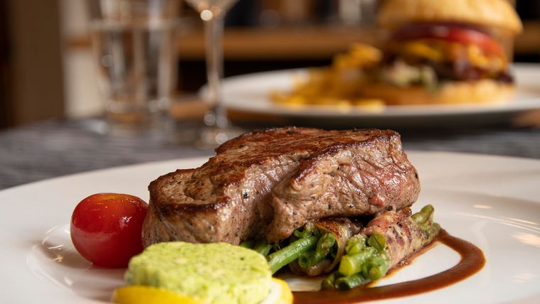 Ein saftiges Steak mit grünen Bohnen, einer Zitronenscheibe und einer grünen Sauce. Im Hintergrund ist ein Burger und Getränke zu sehen.