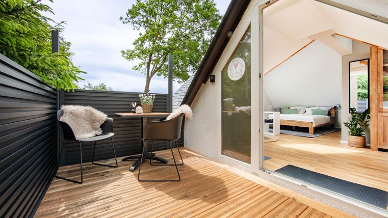 Eine moderne Terrasse mit Holzbelag und zwei Stühlen. Im Hintergrund ist ein helles Zimmer mit einem Bett und Pflanzen sichtbar.