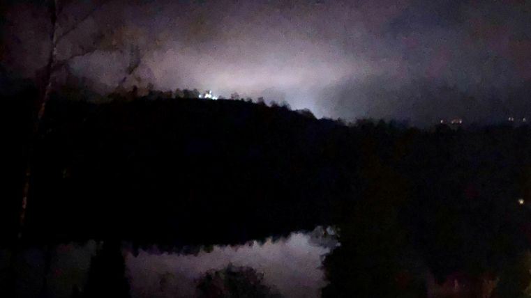 Eine dunkle Nachtlandschaft mit einem ruhigen See und leichten Nebelschwaden. Im Hintergrund sind schwache Lichter zu erkennen.