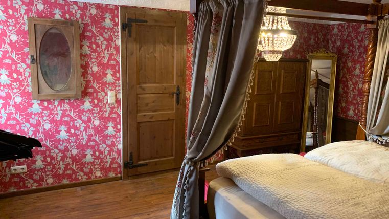Ein gemütliches Schlafzimmer mit einem Himmelbett und einer eleganten Deckenleuchte. Die Wände sind mit blumenmusterndem Tapeten verziert, und es gibt Holzdetails im Raum.