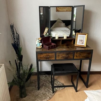 Ein stilvoller Schminktisch mit einem großen Spiegel und einer Blumendekoration. Neben dem Tisch steht ein Hocker und ein Bildrahmen.