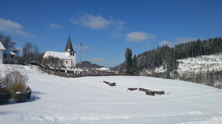 Eine verschneite Landschaft mit einem kleinen, weißen Dorf und einer Kirche im Hintergrund. Der Himmel ist klar und blau, und Bäume sind mit Schnee bedeckt.