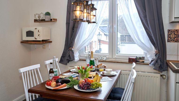 Ein gemütlicher Essbereich mit einem Holztisch und weißen Stühlen. Auf dem Tisch sind verschiedene Lebensmittel und Getränke angerichtet, während das Licht durch die Fenster fällt.