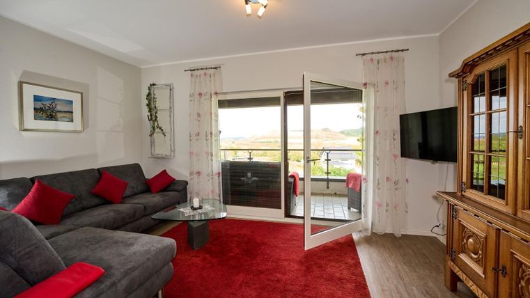 Ein modernes Wohnzimmer mit einem grauen Sofa und roten Kissen. Große Fenster bieten einen Blick auf die Landschaft und der Raum ist hell und einladend.
