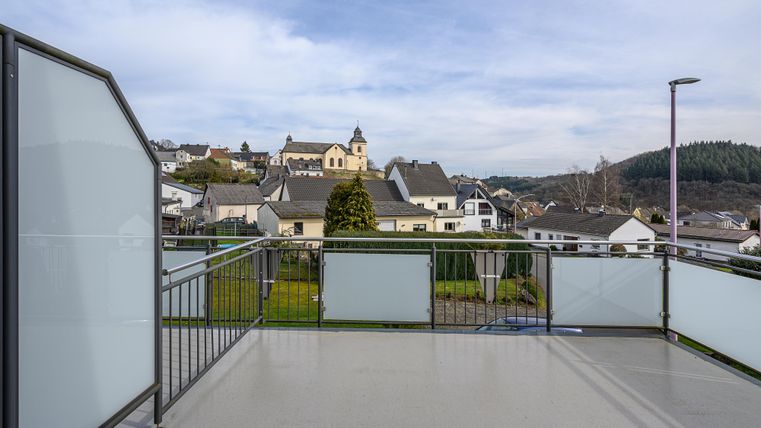 Eine großzügige Terrasse mit Blick auf ein ruhiges Dorf. Im Hintergrund sind Häuser und eine Kirche sichtbar, umgeben von sanften Hügeln.