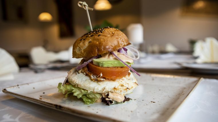 Ein leckerer Burger auf einem Teller, garniert mit Salat, Tomate, Avocado und Zwiebeln. Der Burger hat ein frisches Brötchen und wird in einem stilvollen Restaurant serviert.