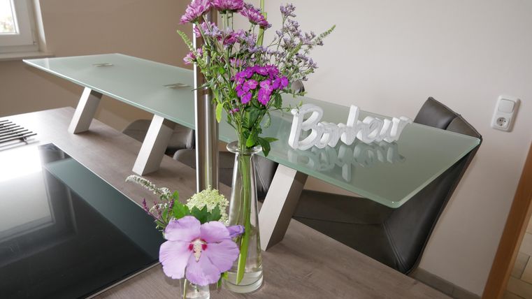Ein moderner Essbereich mit einem gläsernen Tisch und stilvollen Stühlen. Frische Blumen in Vasen und ein dekoratives Schild ergänzen die gemütliche Atmosphäre.
