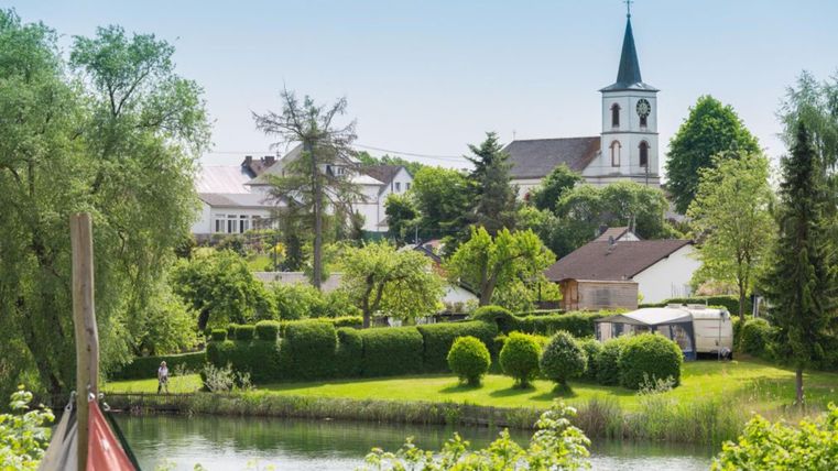 Eine malerische Landschaft mit einem ruhigen Teich und gepflegten Gärten. Im Hintergrund ist eine Kirche mit einem hohen Turm zu sehen.