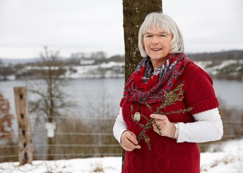 Eine Frau mit grauem Haar steht vor einem See, umgeben von winterlicher Landschaft. Sie trägt einen roten Pullover und hält einen Zweig in der Hand.