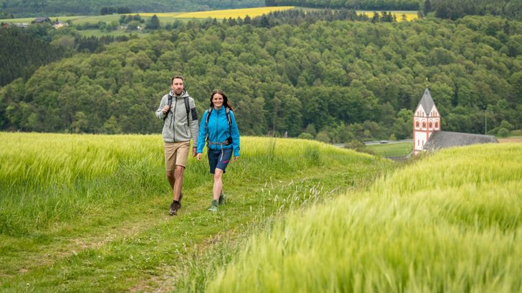 Two hikers on a path through green fields with a church in the background.