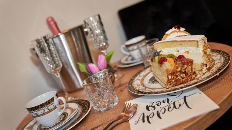 Ein Tischgedeck mit einer köstlichen Torte, elegantem Geschirr und Sektgläsern. Eine kleine Blumendekoration und eine Serviette mit dem Schriftzug „Bon Appétit“ runden das Bild ab.