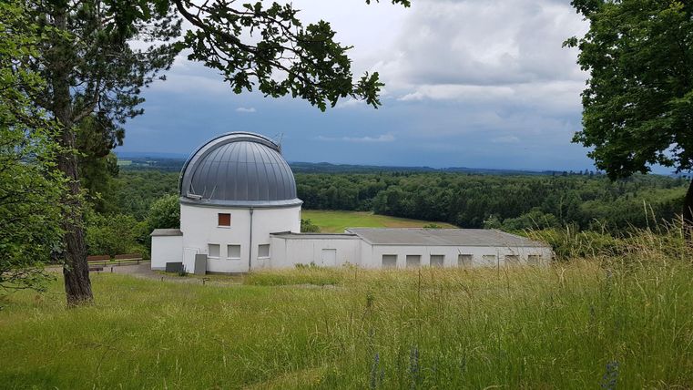 An observatory building amid green meadows and trees. The sky is cloudy, and the landscape stretches over gentle hills.