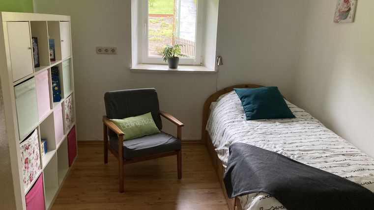 Ein einfaches Schlafzimmer mit einem Bett, einem Stuhl und einem Regal. Das Fenster lässt viel Tageslicht herein und es gibt eine kleine Pflanze auf der Fensterbank.
