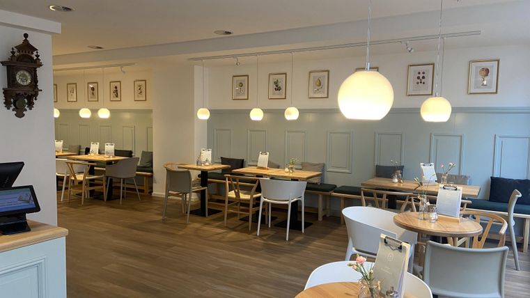 Ein modernes, gemütliches Restaurant mit hellen Tischen und Stühlen. Die Wände sind mit Bildern dekoriert und es gibt sanfte Beleuchtung.