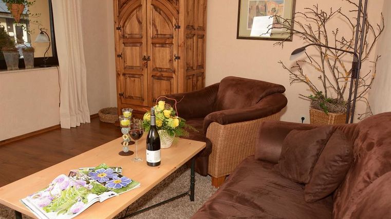 Ein gemütliches Wohnzimmer mit einer dunklen Couch und einem Korbsessel. Auf dem Tisch stehen eine Flasche Wein und frische Blumen.