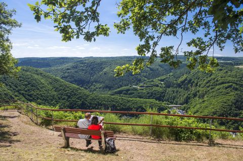 Ein Paar sitzt auf einer Bank und genießt die Aussicht auf das grüne Tal. Der Himmel ist blau und die Landschaft ist üppig und hügelig.