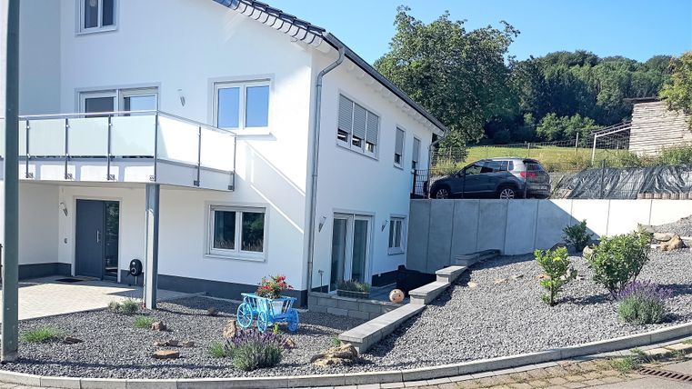 Ein modernes Haus mit einem gepflegten Garten und Kiesfläche. Ein Auto steht in der Einfahrt, und im Hintergrund sind Bäume zu sehen.
