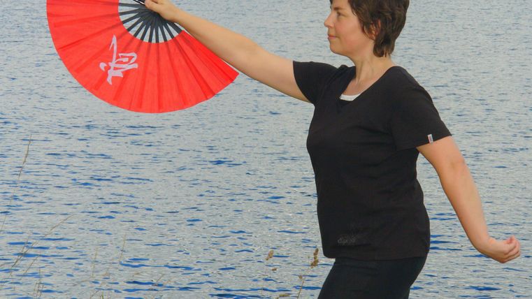 Eine Frau mit einem roten Fächer steht am Ufer eines Gewässers. Sie bewegt den Fächer elegant in der Luft.