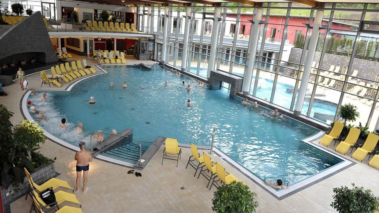 Ein mittlerweile moderner Innenpool mit blauen Wasser und gelben Liegen. Durch große Fenster strömt viel Tageslicht in den Raum.