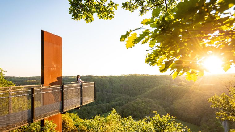 Eine Aussichtplattform in der Natur, umgeben von Bäumen und Landschaft. Die Sonne geht hinter den Hügeln auf und sorgt für eine warme, goldene Atmosphäre.