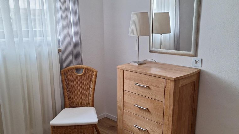 Ein heller Raum mit einem Holzschrank und einem Spiegel. Neben dem Schrank steht ein Stuhl und eine Lampe.