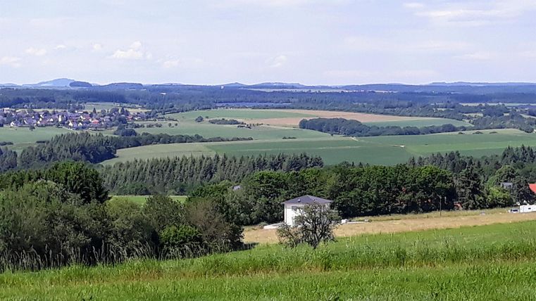 Eine weite Landschaft mit grünen Feldern und Hügeln. Im Vordergrund steht ein kleines Gebäude, umgeben von Bäumen und Wiesen.